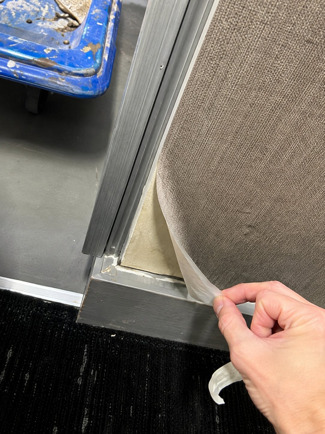 Hand pulling up corner of beige carpet showing gray concrete floor and aluminum trim edge